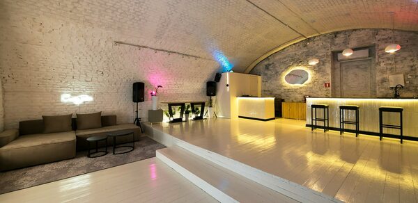Brodniki Loft (Бродники Лофт). Лофт из белого кирпича. Арочный зал с белыми кирпичными стенами и полом. Слева коричневый диван и мягкие кресла на ковре. Справа барная стойка с тремя высокими стульями. Цветное освещение, аудиоколонки. Стены со светлыми и темными оттенками.