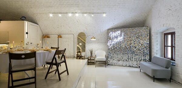 Brodniki Loft (Бродники Лофт). Лофт из белого кирпича. Арочное пространство с белыми кирпичными стенами, обеденной зоной с накрытым столом и стульями, зеркальной стеной с неоновой надписью "Happy Birthday". Деревянный пол, светлые цвета, мягкий серый диван, подвесные светильники.