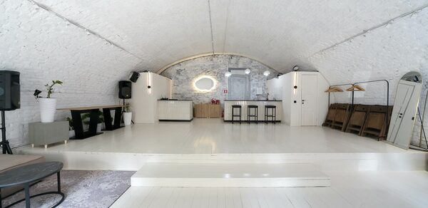 Brodniki Loft (Бродники Лофт). Лофт из белого кирпича. Арочное помещение белого цвета с деревянным полом и ступенькой. На заднем плане барная стойка, стулья, колонки и акустическое оборудование. Декоративное зеркало на кирпичной стене. Слева стулья и журнальный столик. Освещение встроенное.