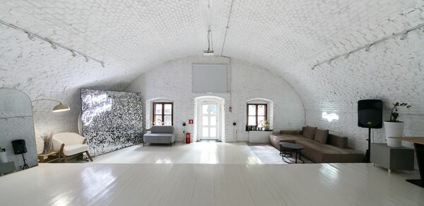 Brodniki Loft (Бродники Лофт). Лофт из белого кирпича. Белый сводчатый зал с деревянным полом, мягкие серые и коричневые диваны, напольная лампа, стена с надписью, три окна, дверной проем, пожарный щит. Потолочное освещение и подвесная небольшая лампа. Стены и потолок из кирпича, белый цвет.