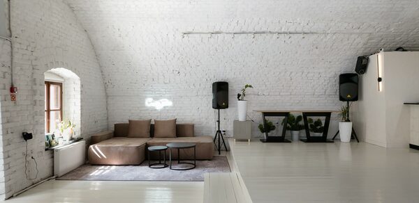Brodniki Loft (Бродники Лофт). Лофт из белого кирпича. Арочное пространство с белыми кирпичными стенами и светлым деревянным полом. Коричневый диван с подушками на ковре, рядом с круглым столиком. По обеим сторонам украшения в виде растений в белых горшках. Два черных динамика. Окно с белыми рамами слева.