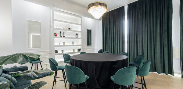 Rubin Loft (Рубин Лофт). Зал Империал. Просторная комната с круглым столом, покрытым черной скатертью, окруженным зелеными стульями. На стене белые полки с декором, в центре потолка большая люстра, длинные темные шторы, плиточный пол, комнатные растения.
