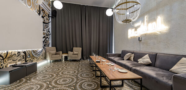 Rubin Loft (Рубин Лофт). Зал Дубай. Зал с диваном и креслами, длинным деревянным столом и потолочными люстрами. Стены украшены узорами, на одной из стен неоновая надпись. На полу ковер с геометрическим узором. Большие темные шторы закрывают окно.