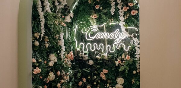 Лофт Кэнди. Арка с декором из зелени и цветов, неоновая вывеска с текстом "Candy" на зелёной стене с растениями. Цветы белые и розовые, стенка покрыта плотной листвой. Освещение от вывески; рамка арки светлого оттенка.