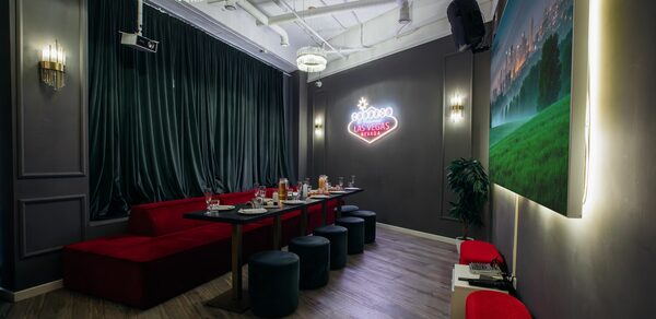 Rubin Loft (Рубин Лофт). Лофт Вегас. Караоке-зал с серыми стенами и деревянным полом. Длинный стол с черными стульями и красными диванами. Верхний неоновый знак "Welcome Las Vegas". Большая картина на стене, темные шторы. Потолочные светильники и проектор.