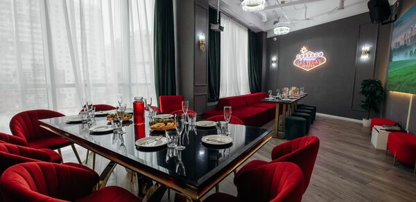 Rubin Loft (Рубин Лофт). Лофт Вегас. Современное помещение с застекленными окнами, красные кресла вокруг длинного стола с черной глянцевой поверхностью, сервировка столовых приборов, диваны у стены, неоновая надпись, люстры, деревянный пол, темные шторы, зеркало на стене.