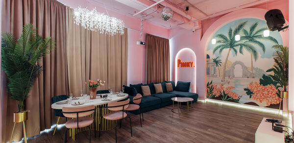 Rubin Loft (Рубин Лофт). Лофт Пинки. Праздничное пространство с розовыми стенами, деревянным полом. Угловой синий диван, круглый обеденный стол с розовыми сиденьями. Подвесной светильник, зеркало в форме арки с подсветкой. Настенный декор с тропическим рисунком, растение в горшке.