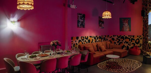 Rubin Loft (Рубин Лофт). Лофт Гламур. Розовая комната с обеденным столом и стульями, сервировка на столе, оранжевый угловой диван, круглый столик, декор с леопардовым принтом, настенные картинки, подвесные светильники с абажурами, светлое точечное освещение, пол темного цвета.