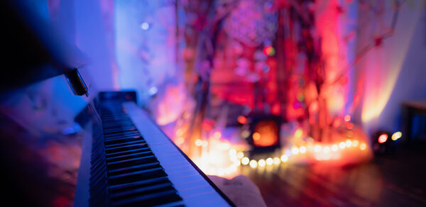 Лес. Театральный лофт с верандой, сценой и вторым ярусом. Пианино крупным планом с боковой стороны, на заднем плане декор из веток с цветными огнями и гирляндами. Мягкое разноцветное освещение в синих и красных тонах. Пол тёмного оттенка, стены светлые.