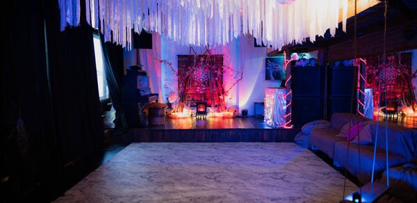 Лес. Театральный лофт с верандой, сценой и вторым ярусом. Просторное помещение с белым узорчатым полом и темными стенами, украшено длинными белыми лентами под потолком. По периметру разнообразная мебель, включая диван и декорации с разноцветной подсветкой. На сцене красочная инсталляция. Свет приглушен.