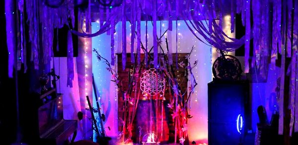 Лес. Театральный лофт с верандой, сценой и вторым ярусом. Темное пространство с ярким разноцветным освещением. Потолок украшен множеством висящих лент и гирлянд. В центре декоративная композиция с ветками и светящимися элементами. Видны мебель и оборудование, такие как фортепиано и аудиоколонки.