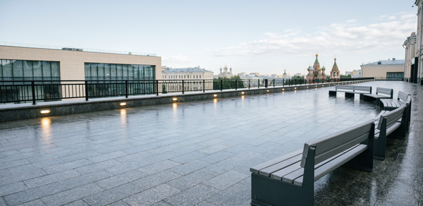 Площадка в Москве. Открытая просторная терраса с видом на город, покрыта светлым гладким камнем. На площадке установлены современные деревянные лавочки. По периметру ограждение с металлическими решетками. Освещение встроено в пол вдоль ограждения.
