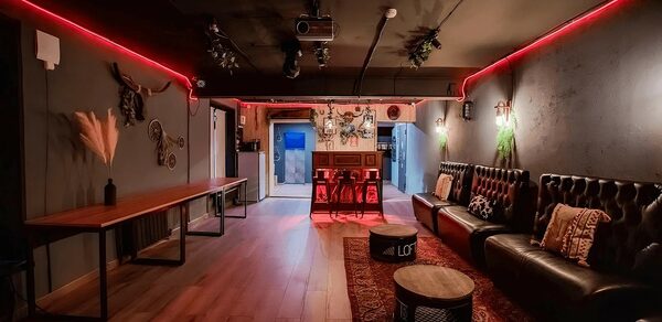 Пространство для любых мероприятий с верандой и грилем. Лофт-пространство с деревянным полом, черной и красной цветовой гаммой. Мебель: длинный деревянный стол, коричневый кожаный диван, круглые столики. Освещение: красные неоновые лампы, настенные светильники. Декор: растения, картины, декоративные головы животных.