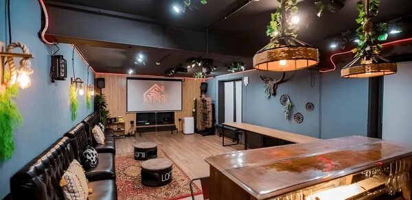 Пространство для любых мероприятий с верандой и грилем. Просторное помещение с мягкими диванами вдоль стены, барной стойкой и длинным деревянным столом. Настенное освещение в виде ламп, подвесные светильники с растениями, экран с проектором. Декор из зелени и светлое деревянное покрытие пола. Стены синие.