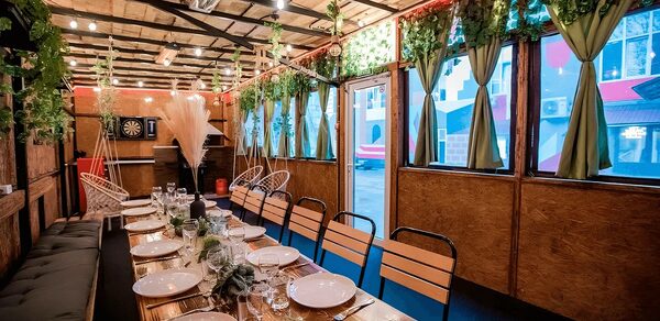 Пространство для любых мероприятий с верандой и грилем. Длинный обеденный стол с белыми тарелками и стеклянной посудой, деревянные стулья, свисающие растения, зелёные шторы на окнах, деревянные панели, подвесные светильники, имитация травы на потолке, дартс на стене, уютный интерьер.