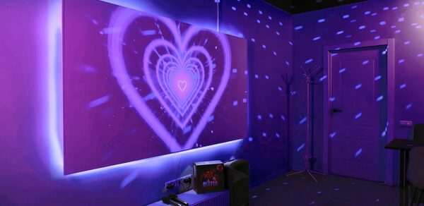 Лофт-зал с караоке и диско-шаром. Комната с фиолетовой светодиодной подсветкой на стене, имеющей изображение сердца. На полу зеркало, рядом стоят видеопроектор и колонка. Справа дверь, в углу вешалка для одежды. Стены однотонные, потолок черный. Световые эффекты создают дискотечную атмосферу.