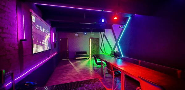 Kahovka loft (Каховка Лофт). Неоновый лофт-зал. Караоке-комната с длинным деревянным столом, стульями, экраном на стене. Стены и потолок украшены светодиодными лентами фиолетового и зеленого цветов. На полу плитка и паркет. Яркое разноцветное освещение.