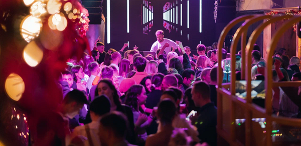 Бар StereoPeople (СтереоПипл). Танцевальная площадка в помещении с диджеем на сцене, окруженной толпой людей. Яркое освещение, верхний светильник и разноцветные декорации. На стене светодиодная панель с геометрическим узором. Справа деревянные конструкции, слева праздничное украшение.
