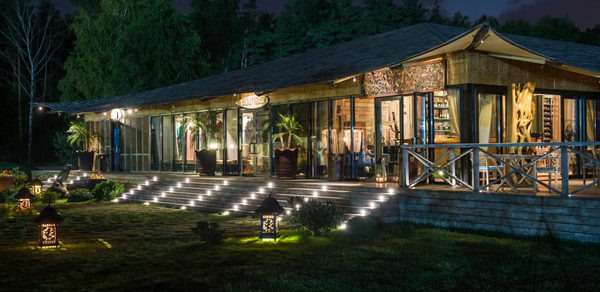 Soho Country Club (Сохо Кантри Клаб). Здание с большой террасой на фоне леса. Стеклянные стены, деревянные конструкции, крыша с вытянутым навесом. Перед входом ступени с боковой подсветкой. На газоне декоративные фонари и растительность. Внутри видны деревянные элементы и растения.