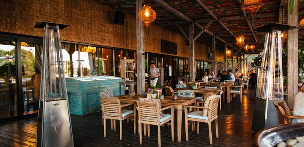 Soho Country Club (Сохо Кантри Клаб). Открытая терраса с деревянными полами и потолком, украшена висящими светильниками. Расставлены столы и стулья из светлого дерева. На заднем плане стеклянные двери, по бокам уличные обогреватели. Декор в теплых тонах с зелеными элементами.