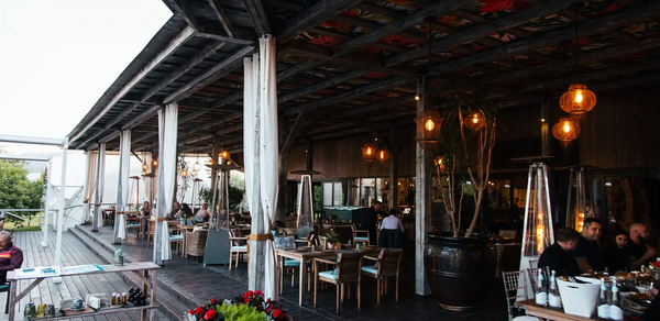 Soho Country Club (Сохо Кантри Клаб). Открытая терраса с деревянным полом и крышей, выбеленными колоннами, множеством деревянных столов и стульев. Подвесные светильники, горшок с растениями и обогреватели. На столах белая посуда. В кадре несколько людей за столами.