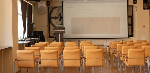 Зал 9. Просторный конференц-зал до 200 человек в Академии Кантаты. Конференц-зал с бежевыми стульями в ряд, деревянный пол, проектор с экраном, аудиосистема, кафедра, монитор, светлые стены, окна с занавесками, потолочные светильники.