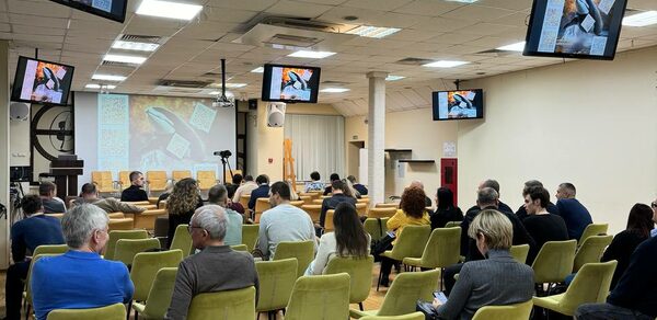 Зал 9. Просторный конференц-зал до 200 человек в Академии Кантаты. Конференц-зал с рядами зеленых стульев, оборудован проектором и несколькими экранами, показывающими изображения. Потолок с встроенными светильниками. Люди сидят, некоторые с блокнотами. Белые стены, деревянный пол, хорошее состояние.