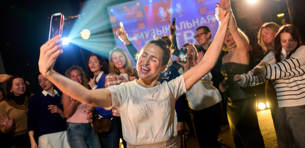 Лофт для мероприятий с гольф-симулятором. Люди танцуют перед проектором с текстом на экране, красочное свечение от прожекторов, множество молодых людей в неформальной одежде, светлые и темные тона в одежде, разнообразные эмоции и движения, вечернее освещение.