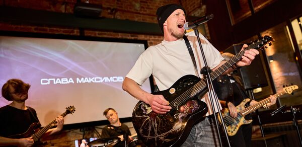 Лофт для мероприятий с гольф-симулятором. Зал с кирпичными стенами, сценой и музыкальными инструментами, включая гитары и барабаны. Мужчина поет в микрофон, держа гитару. На заднем плане экран с текстом. Освещение точечное, теплый свет.