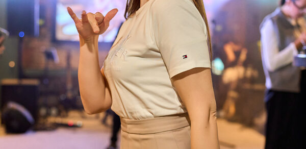 Лофт для мероприятий с гольф-симулятором. Человек в светлой футболке и бежевой юбке в помещении с мягким светом. Видна акустическая система и сцена на фоне. Настенное покрытие — кирпич. Размытый фон с людьми и световыми установками.