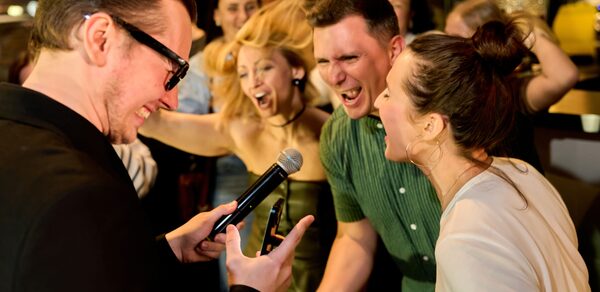 Лофт для мероприятий с гольф-симулятором. Группа людей веселится в помещении, один человек держит микрофон. Видна кирпичная стена, задний план размытый. Яркое освещение, тёплые цвета, присутствует барная стойка.