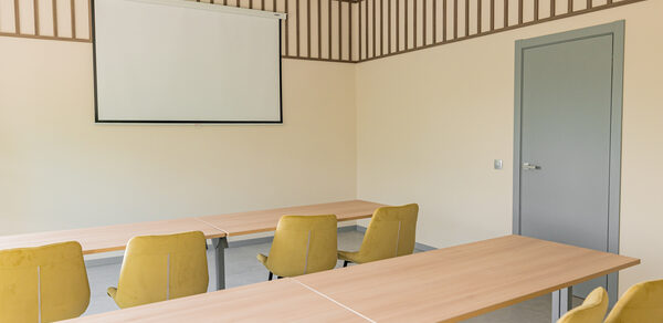 Зал 5. Светлый малый конференц-зал до 20 человек в Академии Кантаты. Просторная комната с бежевыми стенами и потолком с деревянными панелями. В центре расположены столы из светлого дерева и желтые стулья. На стене висит проекционный экран. Справа серая дверь. Естественное освещение.