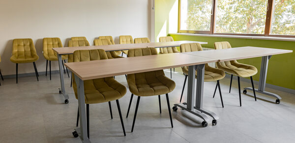 Зал 5. Светлый малый конференц-зал до 20 человек в Академии Кантаты. Конференц-зал с бежевыми стульями и столами на колесиках. Светлая комната с большими окнами, светло-зеленые и белые стены, серый пол. Естественное освещение из окна. Жалюзи частично опущены.