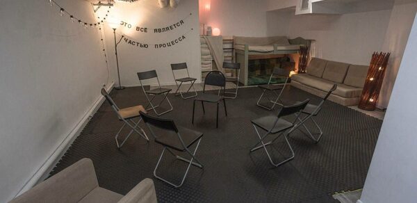 Пространство Нора лофт. Лофт-пространство с серыми стенами и потолком, украшенное гирляндами. По центру расставлены складные стулья, вдоль стены диван и кресло. Черный резиновый пол, лестница ведет на второй уровень. Надпись на стене из черных букв.