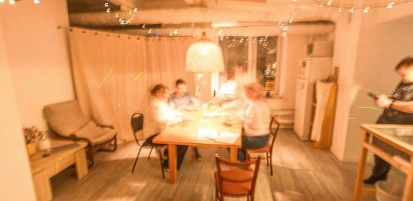 Пространство Нора лофт. Просторная комната с большим деревянным столом и стульями, окруженная занавесками и декорированная подвесными гирляндами. В комнате есть диван и кухня с холодильником. Мягкое освещение от большого подвесного светильника. Пол покрыт плиткой.