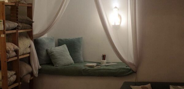 Пространство Нора лофт. Уютное пространство с мягкой зоной для отдыха, обустроенной в нише с подушками и покрывалом зелёного цвета. Справа диван с подушками, слева стеллаж с текстилем. На стене светильник, создающий тёплое освещение. Нейтральные цвета.