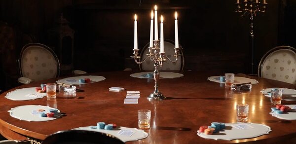 Polygon(Полигон). Зал Апартаменты. Большой круглый деревянный стол с нотками ар-деко, окруженный классическими стульями с узором. На столе расположены канделябры с зажженными свечами, карточки, стаканы и игровые фишки. Освещение теплое, приглушенное. Цвета темные и нейтральные.