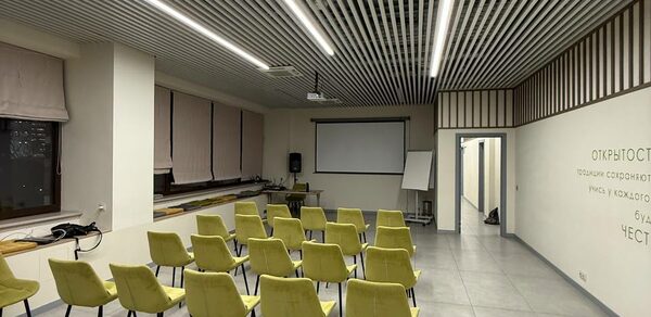 Зал 6. Просторный и светлый конференц-зал до 50 человек в Академии Кантаты. Конференц-зал с рядами желтых стульев, проектором и экраном. Светлые стены с текстом на русском языке, длинные занавески на окнах. Потолок с ламелями и встроенным освещением, пол серый, кафельный. В углу белая доска.