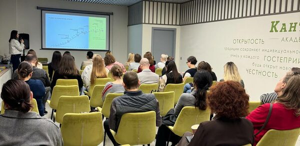 Зал 6. Просторный и светлый конференц-зал до 50 человек в Академии Кантаты. Зал с проектором и экраном, перед которым сидят люди на светло-зеленых стульях. Серые стены с деревянными элементами, на одной из стен текст с крупными словами. Потолок с ламелями, встроенное освещение.