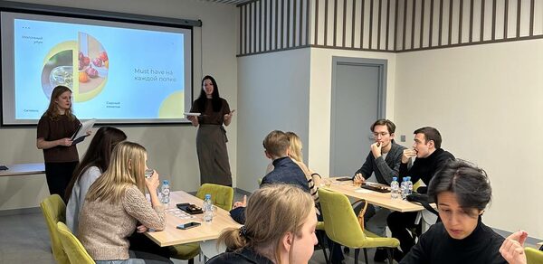 Зал 6. Просторный и светлый конференц-зал до 50 человек в Академии Кантаты. Конференц-зал с тремя длинными деревянными столами и зелеными стульями, светло-серые стены с декоративными полосами, потолок с деревянными панелями. Проектор с экраном, на экране текст и изображение. Несколько людей сидят за столами, двое стоят у экрана.