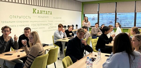 Зал 6. Просторный и светлый конференц-зал до 50 человек в Академии Кантаты. Комната для собраний с современным интерьером, стены украшены текстом, зеленые и бежевые стулья вокруг светлых деревянных столов. Освещение потолочное, большие окна с видом на город. На столах бутылки с водой и чашки.