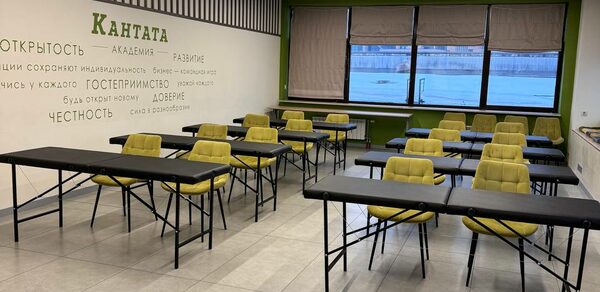 Зал 6. Просторный и светлый конференц-зал до 50 человек в Академии Кантаты. Учебное помещение с черными столами и желтыми стульями, расположенными в рядах. Окно с жалюзи, светлые плиточные полы, зеленая стена с текстом. Потолок с линейными светильниками. Состояние хорошее, естественное и искусственное освещение.