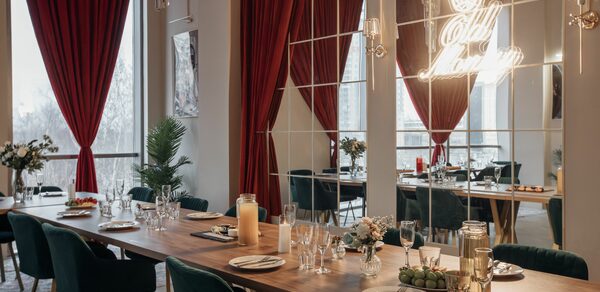 Rubin Loft (Рубин Лофт). Зал Олд Мани. Просторное помещение с длинным деревянным столом, сервированным тарелками и бокалами. Тёмные стулья с мягкой обивкой. Зеркальная стена с подсветкой и надписью. Красные шторы, большие окна, декоративные растения. Мягкое освещение, светлые стены.