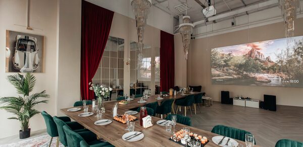Rubin Loft (Рубин Лофт). Зал Олд Мани. Просторное помещение с высоким потолком, длинный обеденный стол с зелёными стульями, сервированная посуда, декоративные элементы, светлый деревянный пол, красные шторы, большие зеркала, проекционный экран, зелёные растения, подвесные люстры.