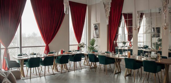Rubin Loft (Рубин Лофт). Зал Олд Мани. Просторное помещение с большими окнами и красными шторами. Длинные деревянные столы с сервировкой и зелёными стульями. Потолочные люстры. Светлые стены и паркетный пол. Зеркала на стене. Справа видна часть дивана.
