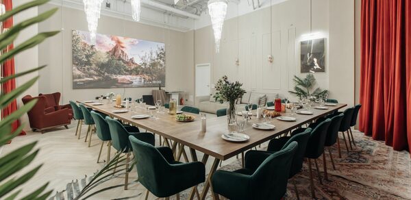Rubin Loft (Рубин Лофт). Зал Олд Мани. Просторное помещение с длинным деревянным столом, окружённым зелёными стульями. Подвесные люстры, красные шторы, ковёр с узором. На стене большая картина и светильник. Растения в горшках, сервировка стола с тарелками и бокалами. Светлые стены и потолок.