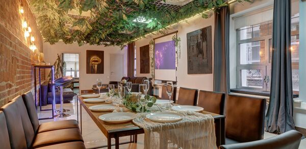 Лофт Семеновский. Праздничное помещение с длинным деревянным столом, окружённым кожаными стульями. На столе белые тарелки и бокалы. Кирпичная стена, занавешенные окна. Потолок украшен зеленью. Мягкое освещение. Окна с видом на город.