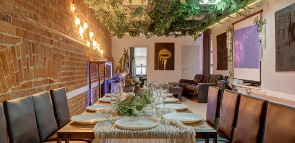 Лофт Семеновский. Лофт-пространство с кирпичной стеной. Длинный стол с сервировкой для гостей, кожаные стулья. Подвесной декор из зелени и гирлянды. На стене проектор с изображением. В зоне отдыха диваны и декор. Потолок металлический.