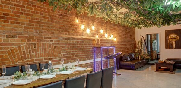 Лофт Семеновский. Пространство с кирпичными стенами и плиточным полом. Длинный деревянный стол с черными стульями, сервирован посудой и бокалами. Потолок украшен зелеными листьями и светодиодными лампами. В углу зона отдыха с кожаными диванами и столиком.