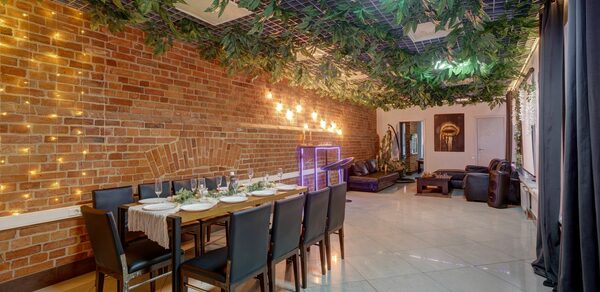 Лофт Семеновский. Праздничное пространство с кирпичной стеной и подвесными растениями на потолке. Стол со стульями сервирован для обеда. В углу мягкая мебель. На стене световые украшения. Плиточный пол, большие окна с черными шторами.